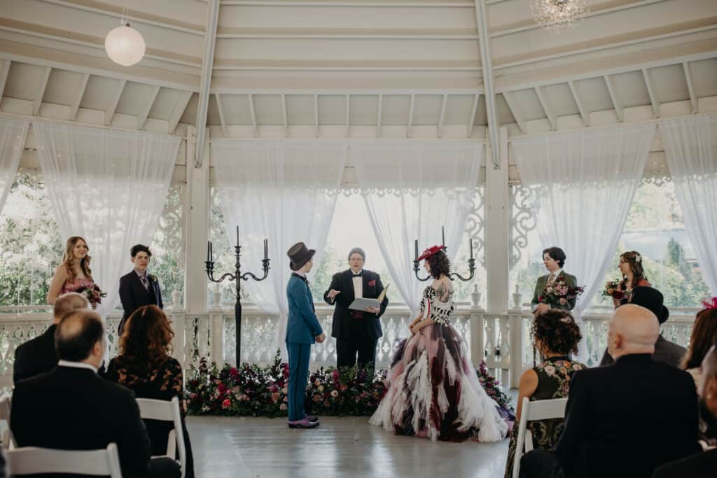 A couple listens to their officiant during a wedding ceremony in the gazebo. Their attendants stand on either side near a large floral display