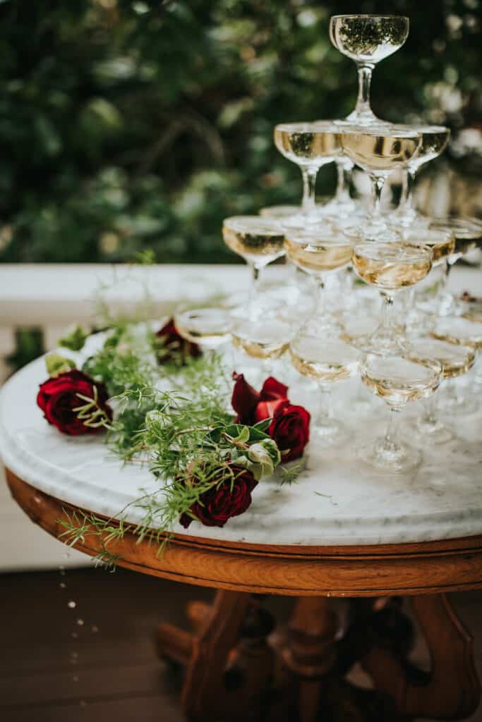 A tower of coupe glasses filled with champagne rests on an antique table decorated with roses.