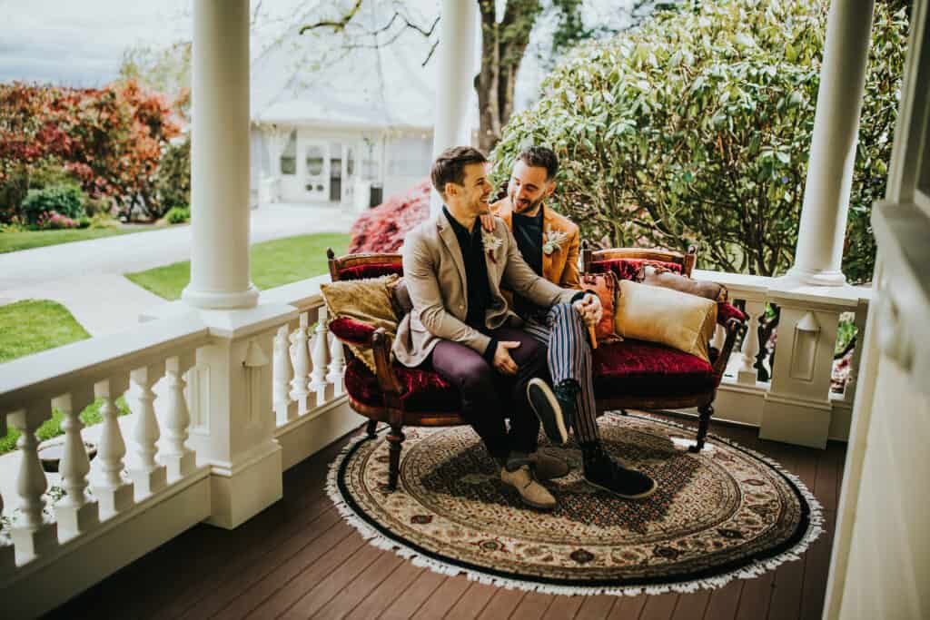A couple sits on an antique velvet couch on the front porch of the Victorian Belle Mansion. Behind them are green and red shrubs and an ornate porch railing