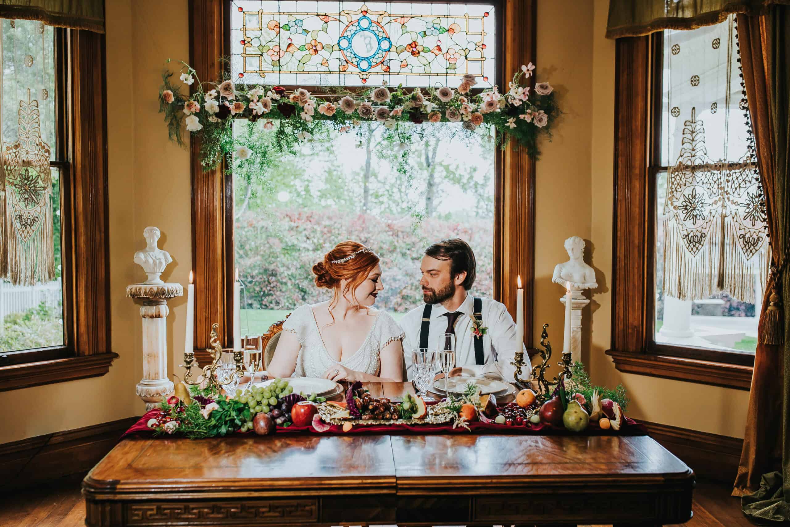 A couple smiles at each other while seated at a table decorated with fruit and flowers. They are seated under stained glass windows with floral accents.