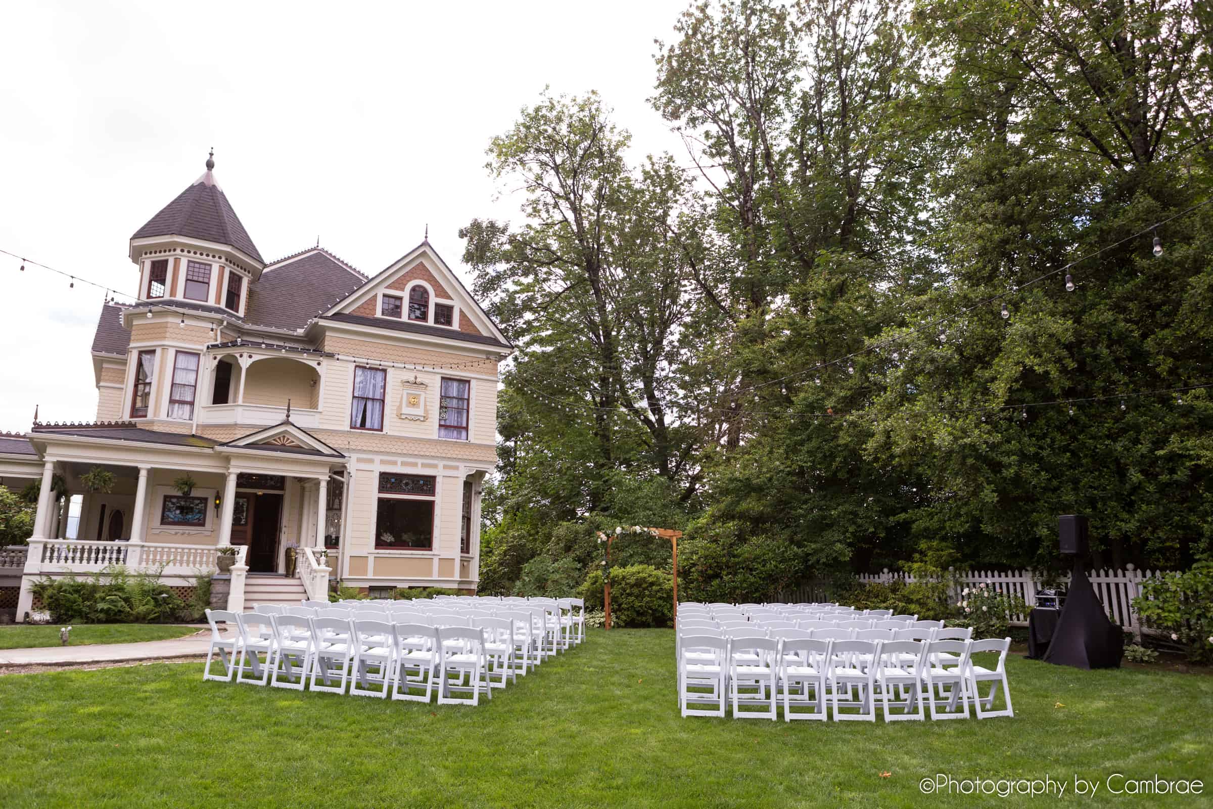 A wedding ceremony with two blocks of chairs and an arbor stand in front of an ornate Victorian mansion.