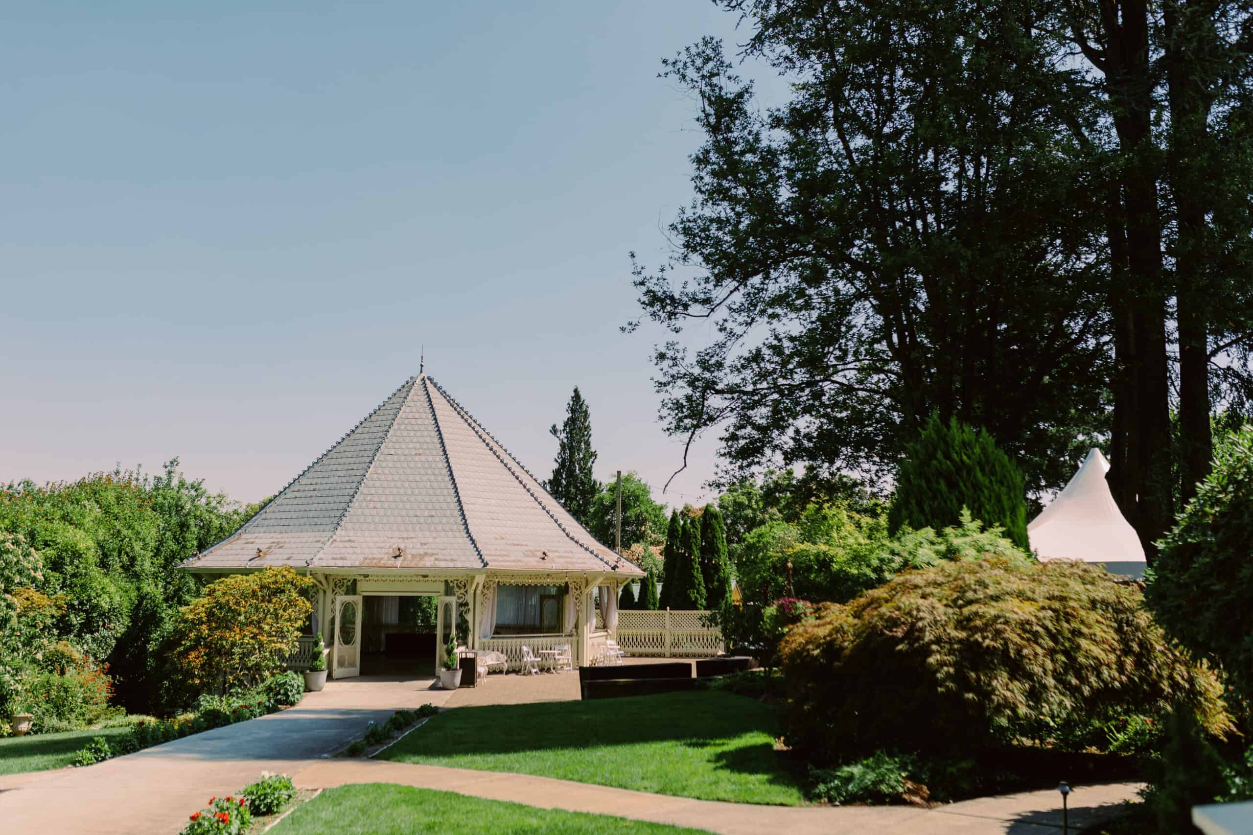 A white and grey gazebo is surrounded by shrubs, lawns, and a courtyard