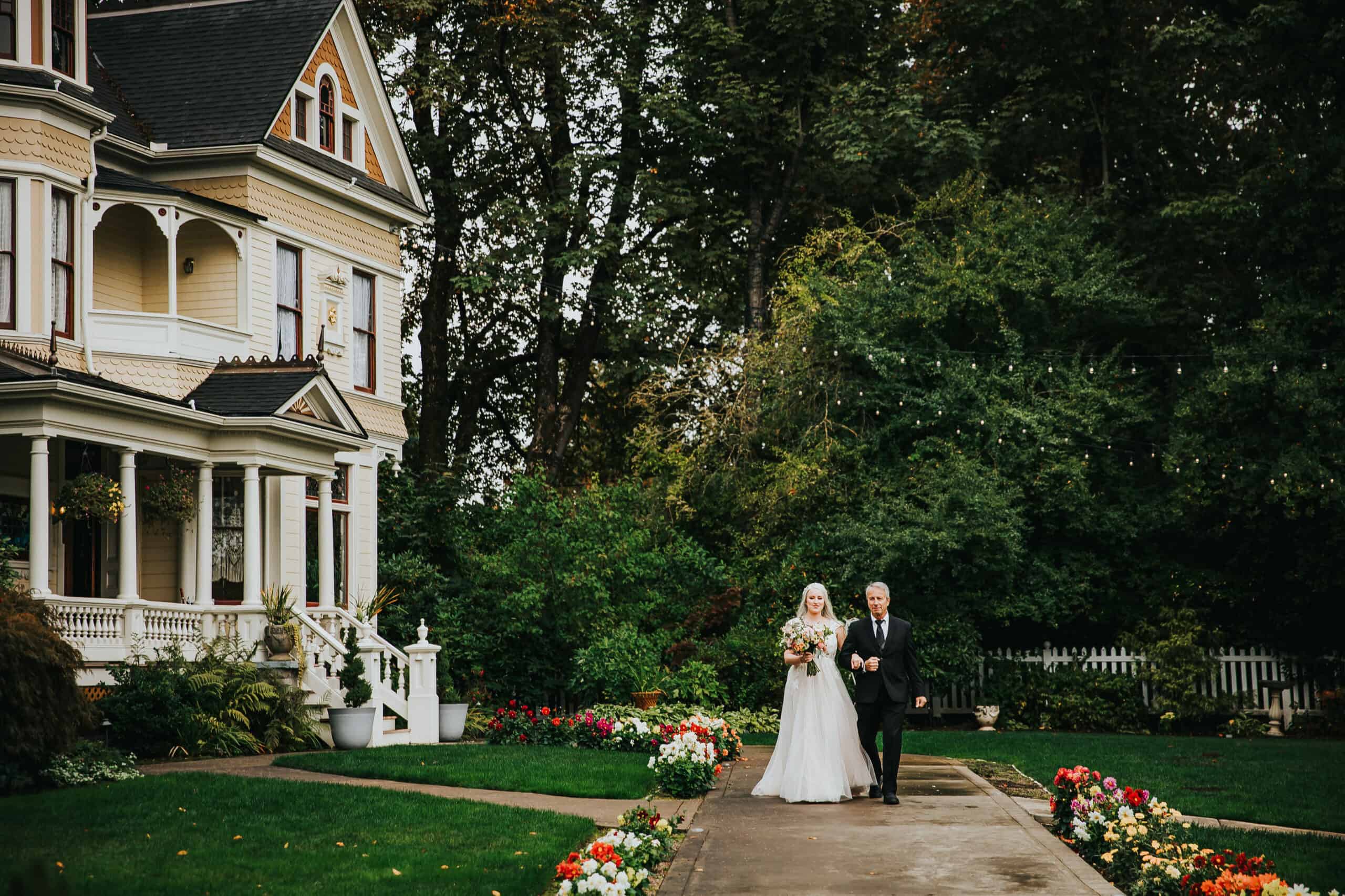 A bride and her father walk down a path away from an ornate Victorian Mansion