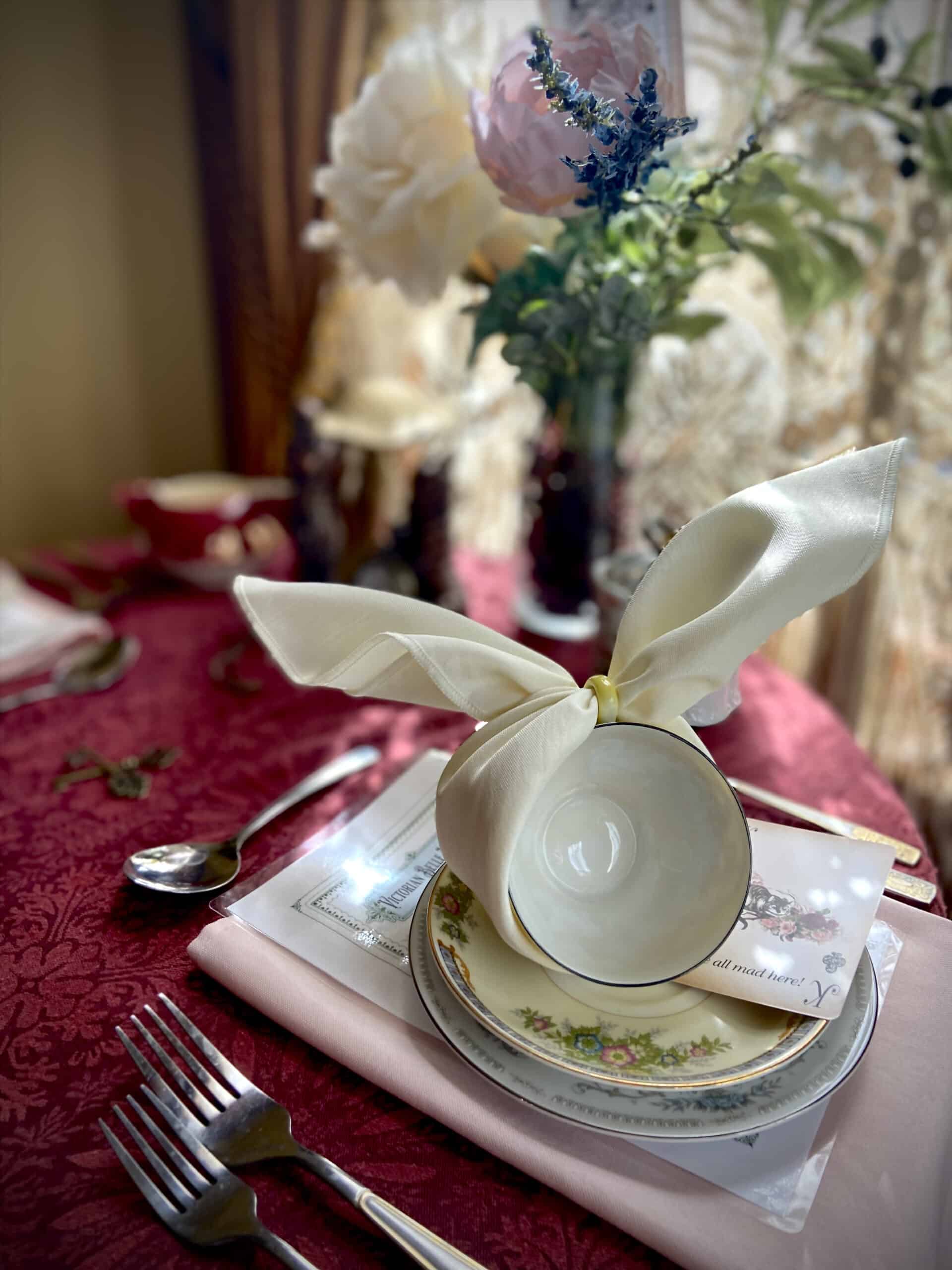 A teacup with napkin tied to look like rabbit ears sits on a plate and saucer with silverware and Alice in Wonderland themed decorations